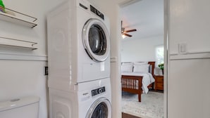 Un espace buanderie avec des machines à laver et des sèche-linge superposés, un lit en bois et un ventilateur de plafond dans une pièce aux murs blancs et au plancher en bois.