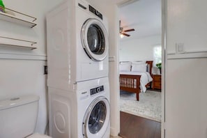 Une buanderie équipée d’une machine à laver et d’un sèche-linge superposés, d’un ventilateur de plafond et donnant sur une chambre avec un lit et une table de chevet.