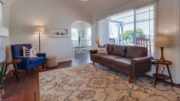 Un salon spacieux avec un canapé marron, un fauteuil bleu et un tapis à motifs. On aperçoit deux lampes et un coin repas au fond de la pièce.