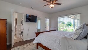 Une chambre à coucher comprenant un lit, un ventilateur de plafond, une télévision, un lave-linge et une fenêtre avec des stores.