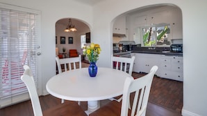 Un coin repas avec une table et des chaises blanches, un vase bleu rempli de fleurs, et une cuisine en arrière-plan.