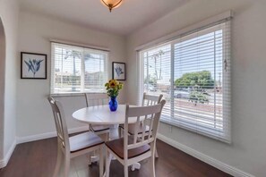 A dining area with a round table and four chairs, two windows with blinds, and a vase with flowers on the table.