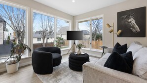 Un salon moderne doté d’une grande fenêtre, d’un canapé noir, d’un pouf rond, d’un lampadaire et d’un téléviseur posé sur un meuble.
