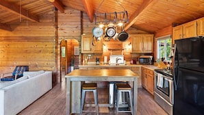 A cozy log cabin interior with a wooden dining table, bar stools, a kitchen with modern appliances, and a comfortable seating area.