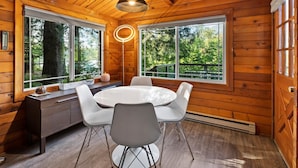 A wooden cabin interior with a round dining table, white chairs, and large windows offering a view of the forest.