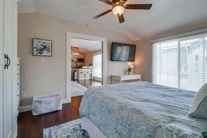 A bedroom with a bed, a ceiling fan, a window with blinds, a door leading to a bathroom, and a small table with a lamp.