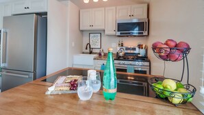A kitchen with white cabinets, a stainless steel refrigerator, a microwave, and a gas stove. There is a fruit basket with apples and a bottle of soda on the counter.