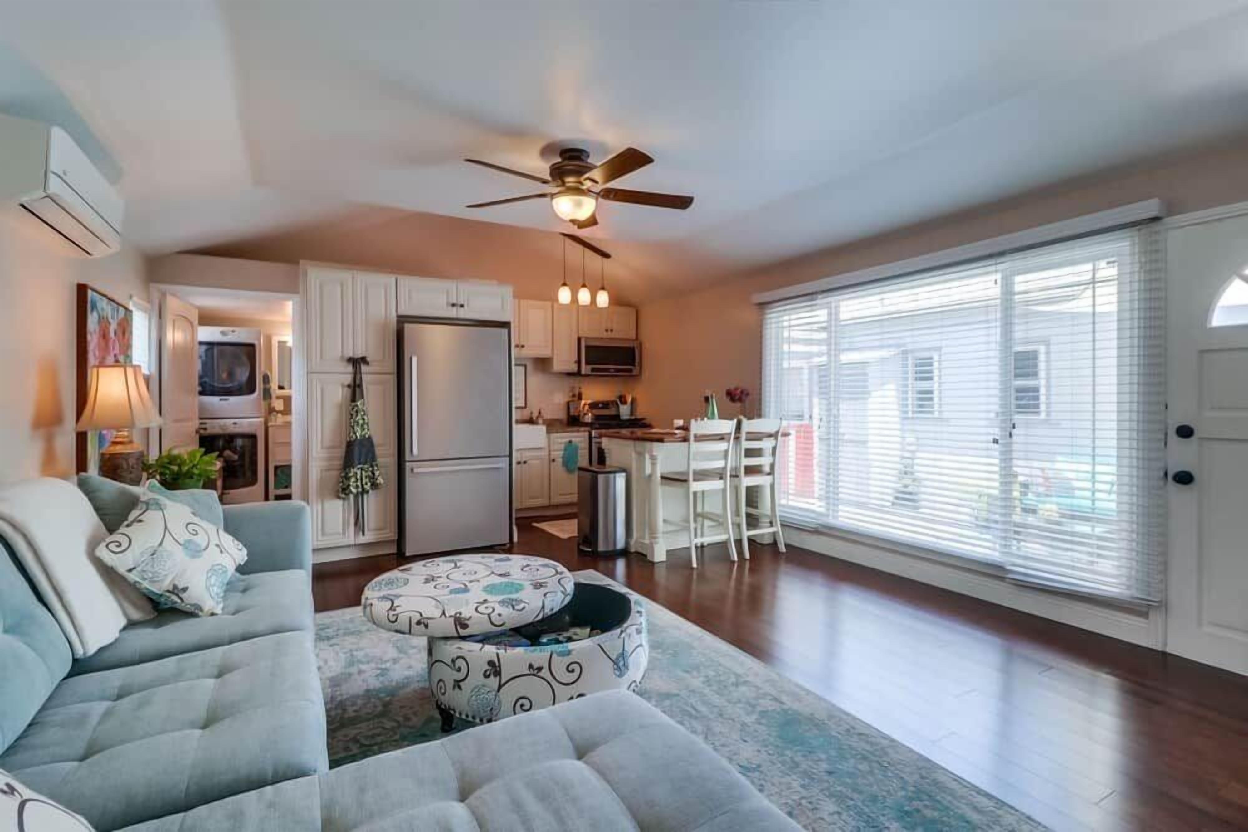 A living room with a sofa, ottoman, and a ceiling fan. There is a kitchen area with cabinets and appliances.