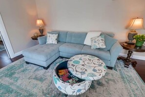 A living room with a light blue sectional sofa, two side tables with lamps, a patterned ottoman, and a rug.