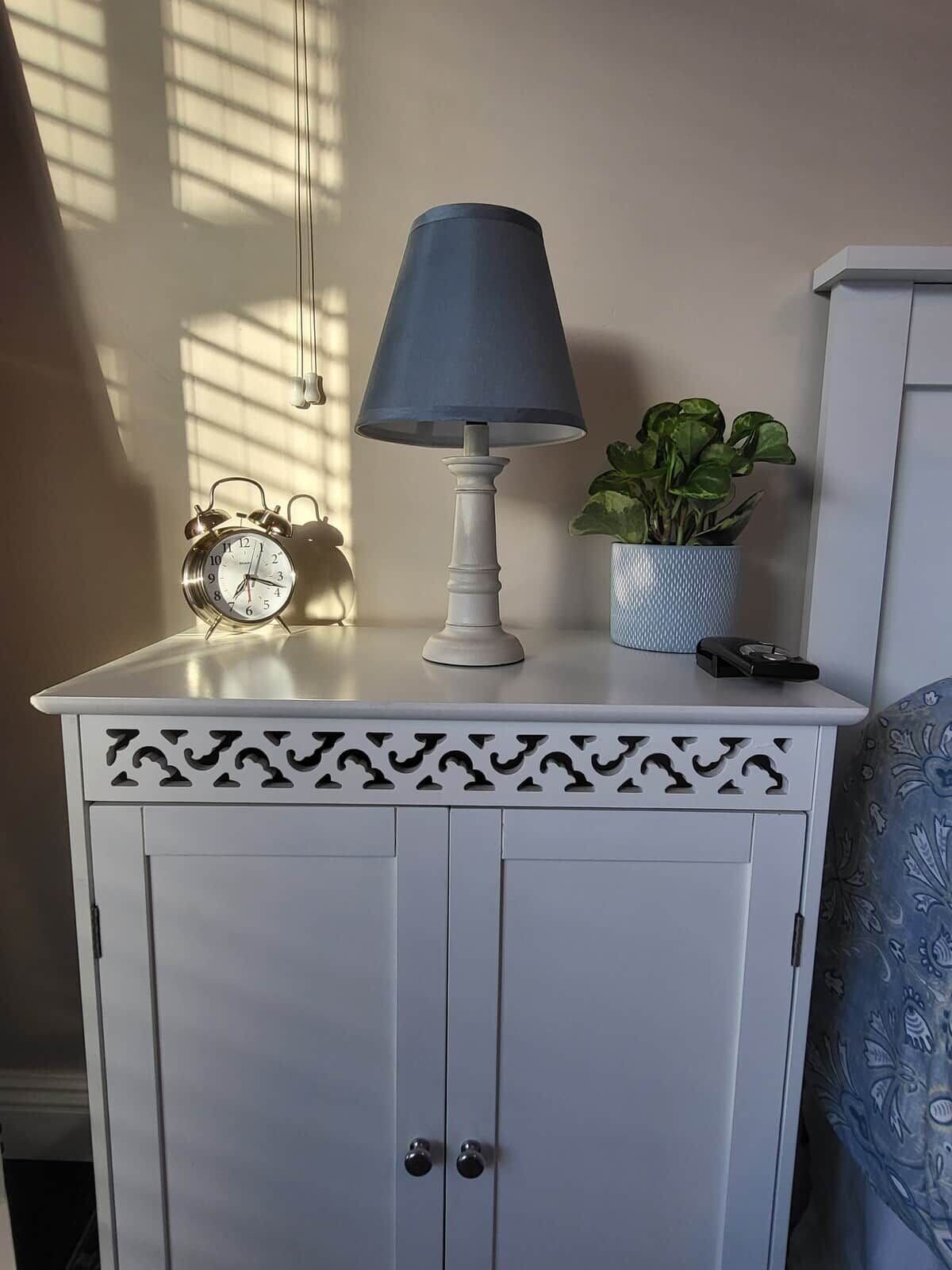 A white bedside table with a blue lamp, a clock, and a plant.