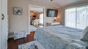 A bedroom with a bed, a ceiling fan, a window with blinds, a door leading to a bathroom, and a small table with a lamp.