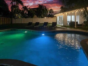 Ein Swimmingpool mit Liegestühlen und einem überdachten Bereich, umgeben von Palmen und einem Sonnenuntergangshimmel.