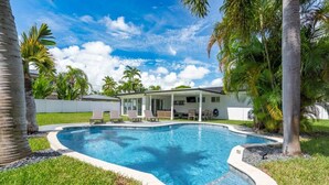 A swimming pool with a curved edge, surrounded by grass and palm trees. - Delila's Chic 3 Bedroom-2 bath w/ Heated Pool + Game Room (Deerfield Beach)