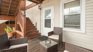 Un espacio exterior techado con mobiliario de mimbre, una mesa de vidrio y una escalera de madera que conduce a una puerta.