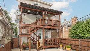 Una casa de dos pisos con terraza de madera, escaleras y patio trasero cercado.