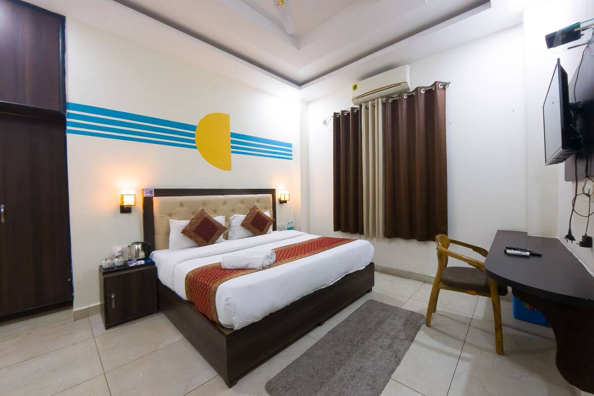 A hotel room with a bed, bedside tables, a desk, a chair, a TV mounted on the wall, and a window with curtains.