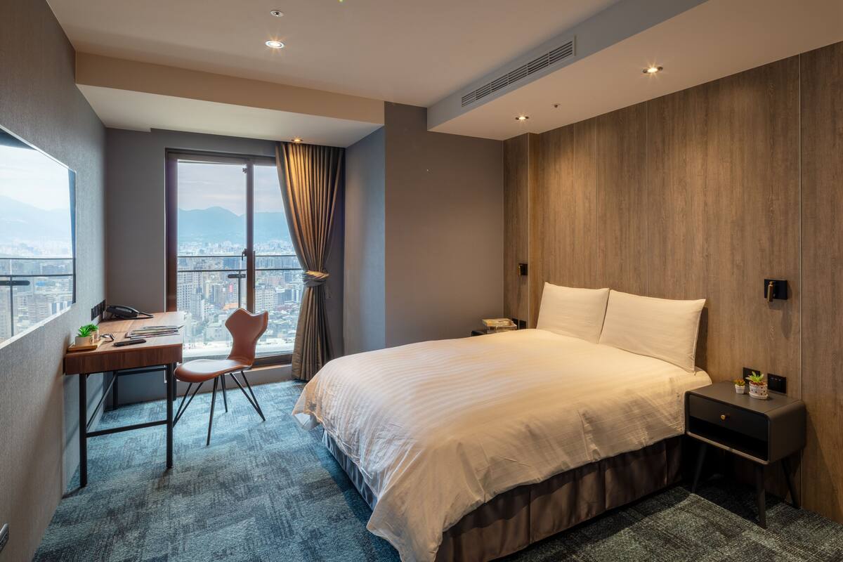 A hotel room with a large bed, a desk with a chair, a window with curtains, and a city view.
