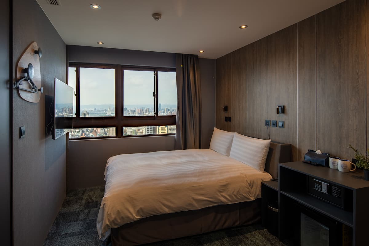 A modern hotel room with a large bed, a city view, a desk with a coffee maker, and a TV mounted on the wall.