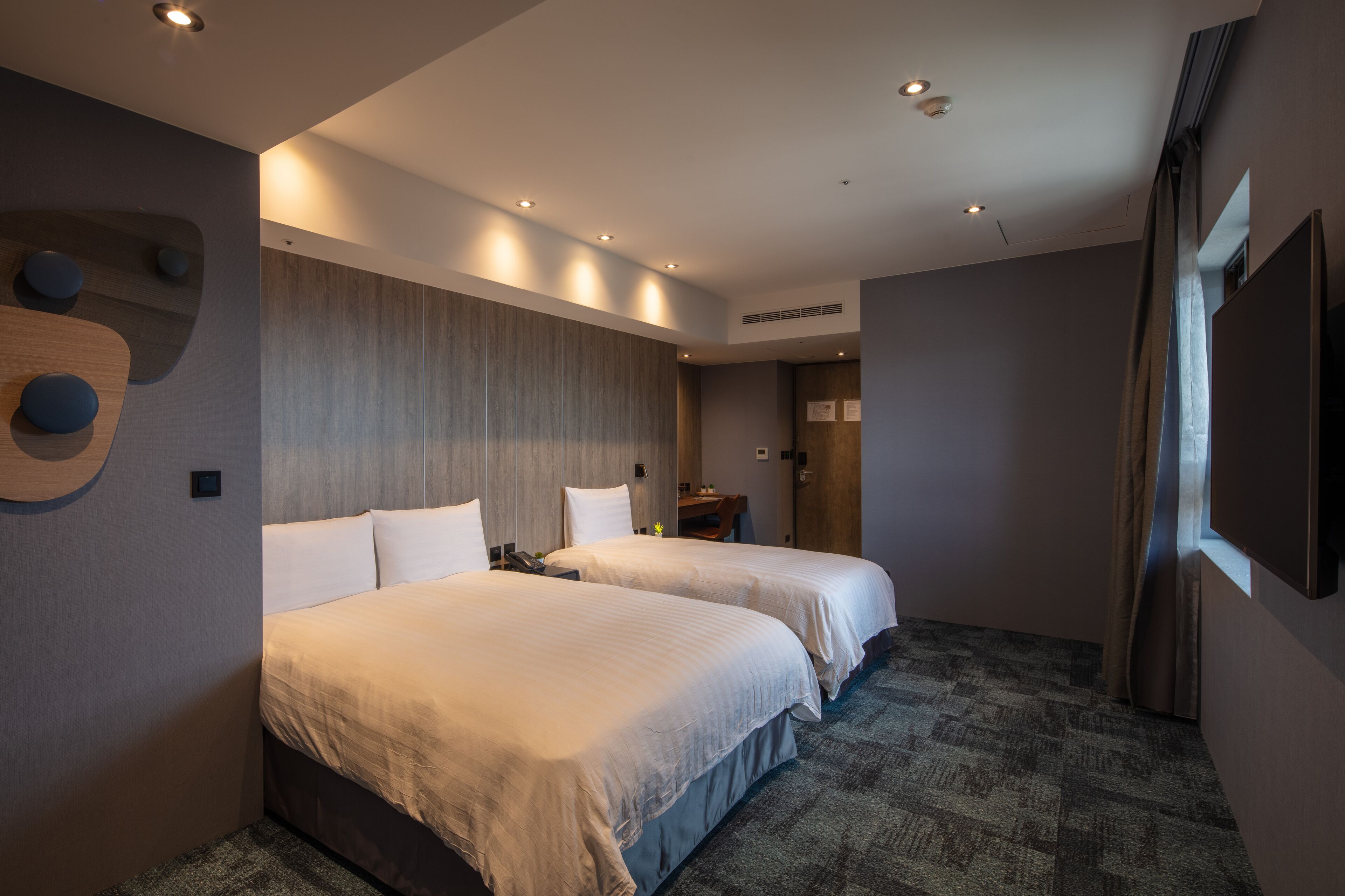 A hotel room with two beds, a wooden headboard, a flat-screen TV, and a small desk with a chair.