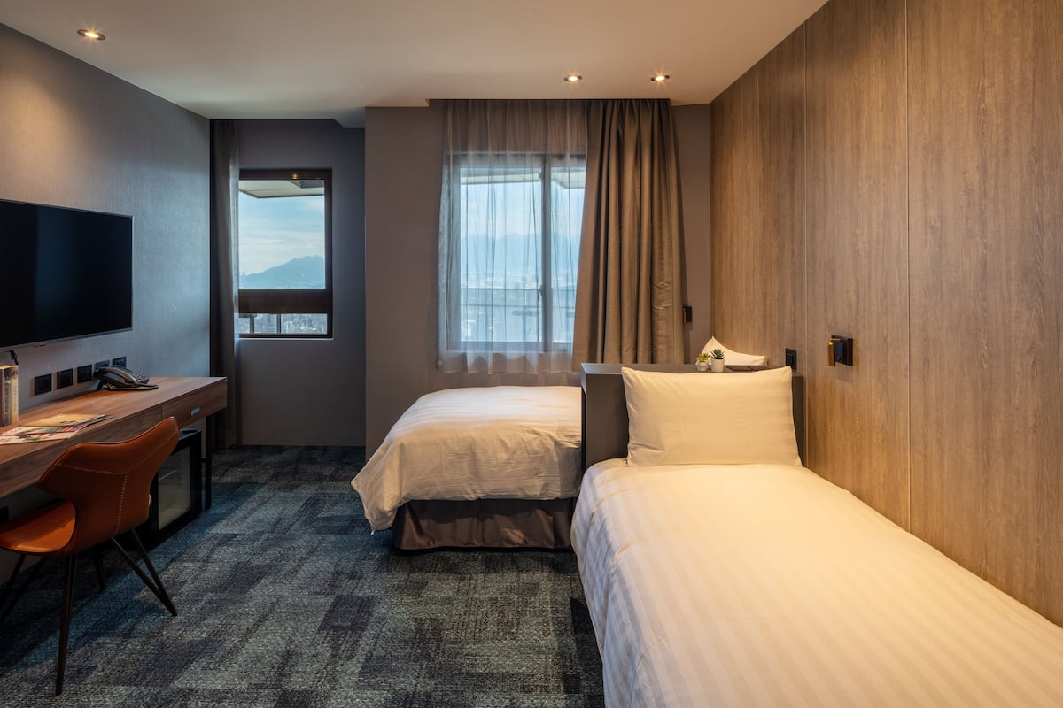 A hotel room with two beds, a desk with a chair, a TV, and a window with curtains.