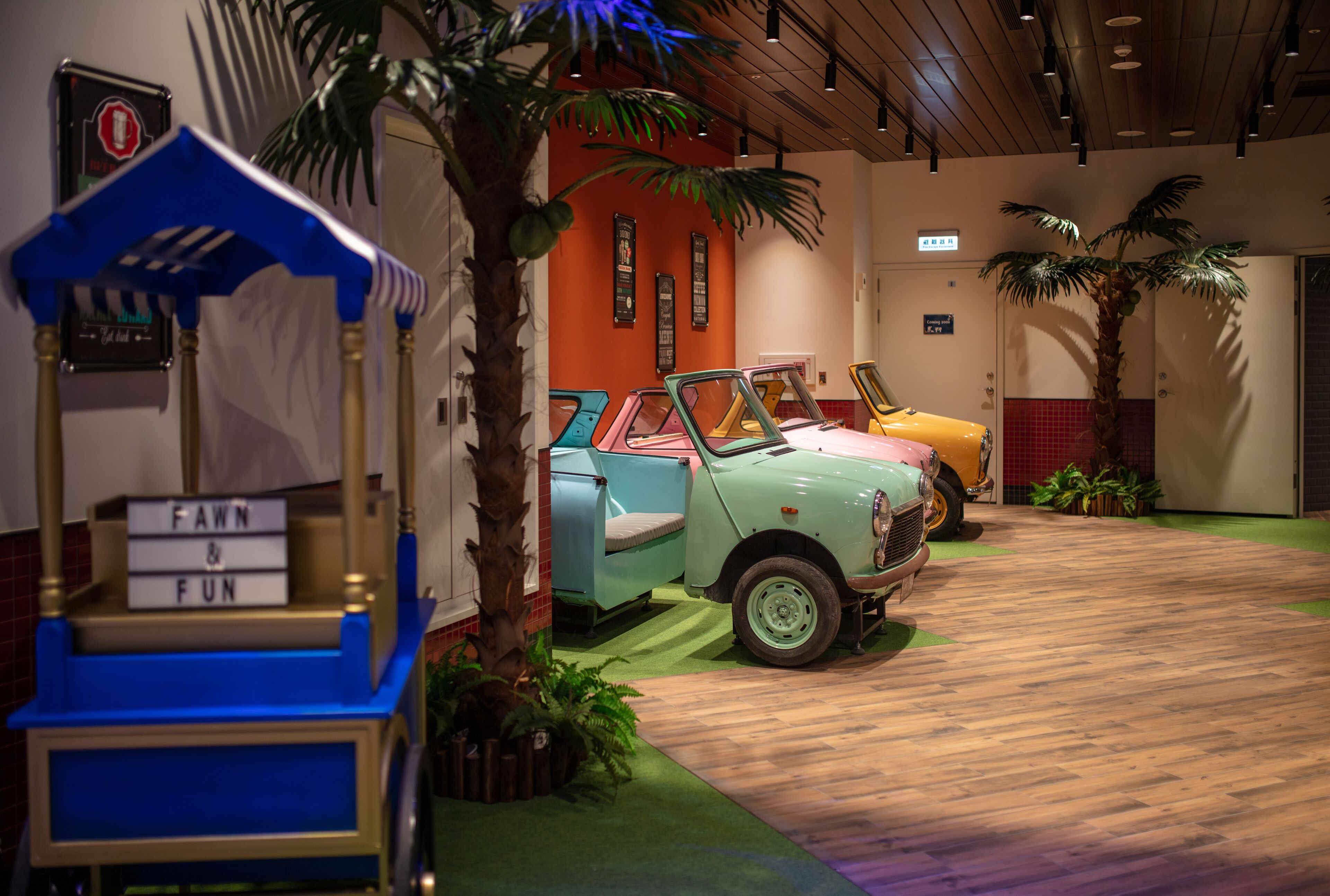A vintage car display with a blue and gold cart labeled "FAWN & FUN" in a room with wooden flooring and tropical plants.