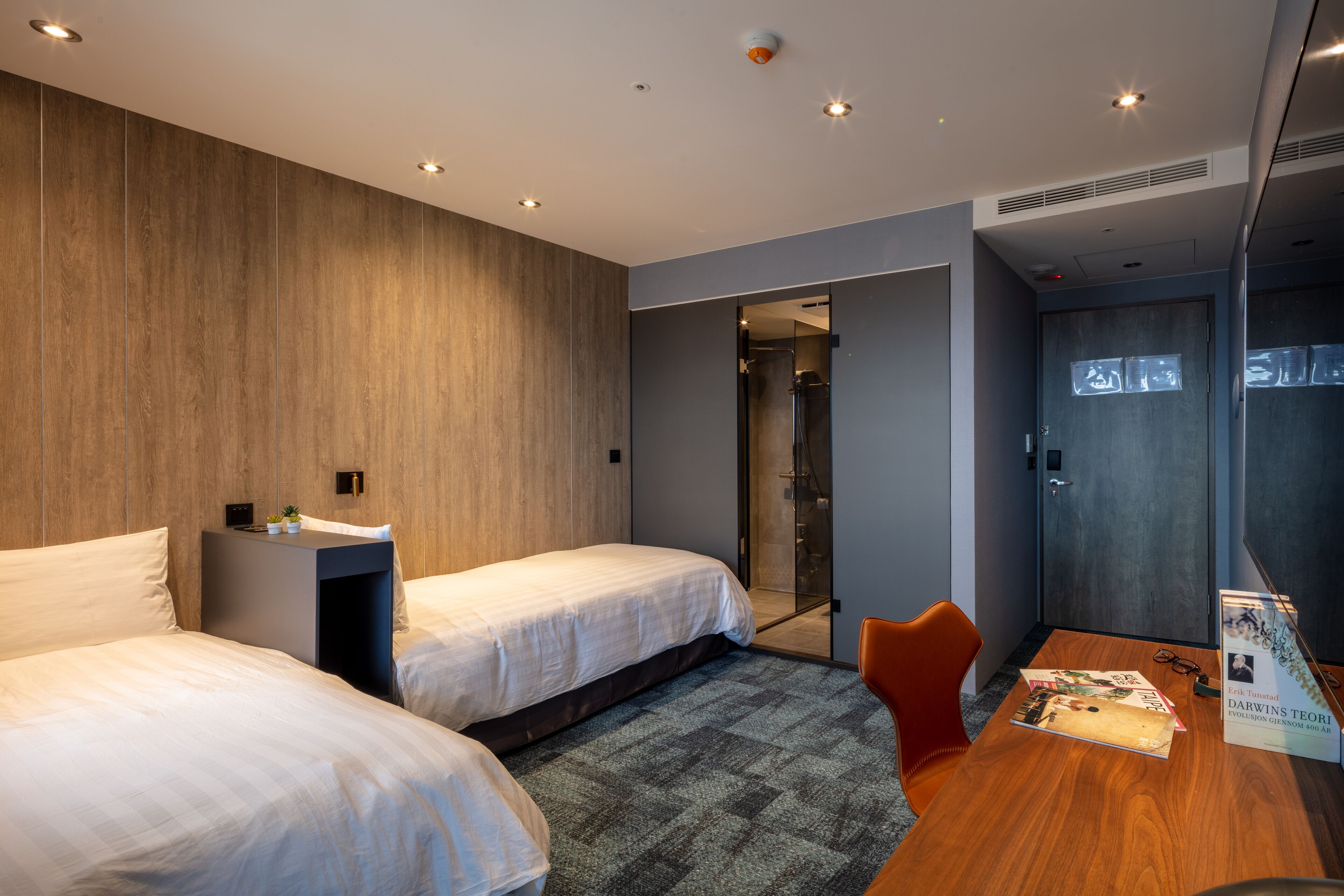 A modern hotel room with two beds, a small table, a chair, and a shower area.
