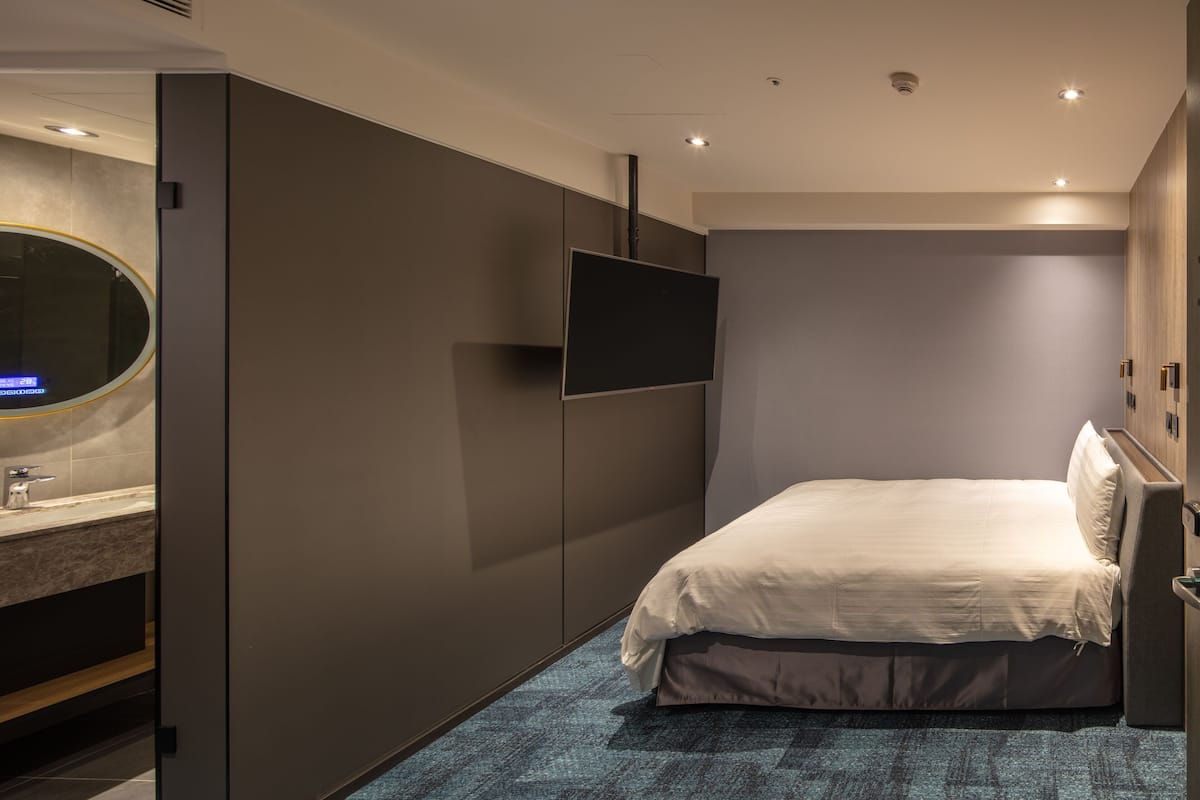 A modern hotel room with a bed, a flat-screen TV mounted on the wall, a bathroom with a mirror and sink, and a blue carpet.