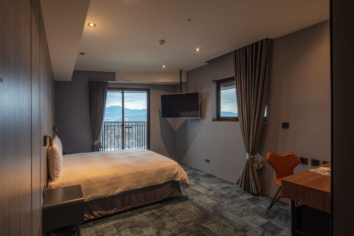 A hotel room with a large bed, a desk, a TV mounted on the wall, and a view of the mountains through the window.