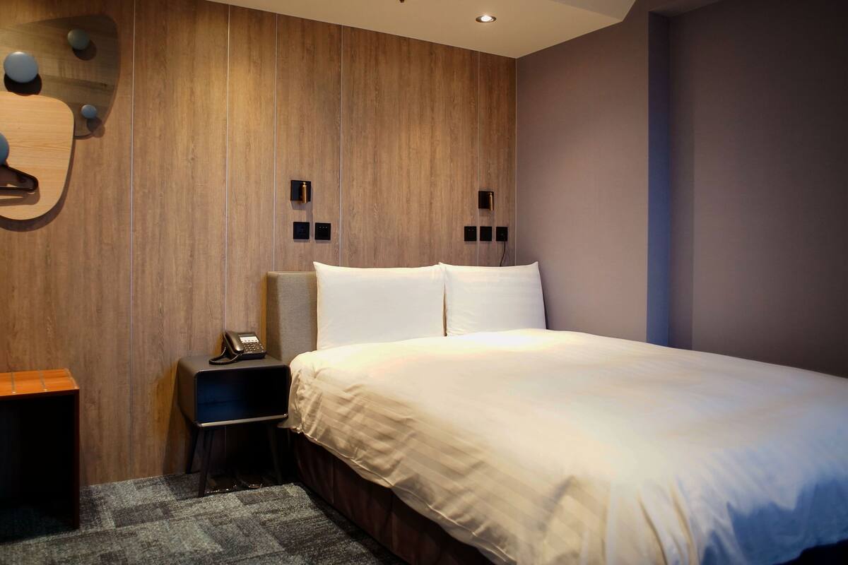 A modern hotel room with a wooden accent wall, a bed with white linens, a nightstand with a phone, and a small table.