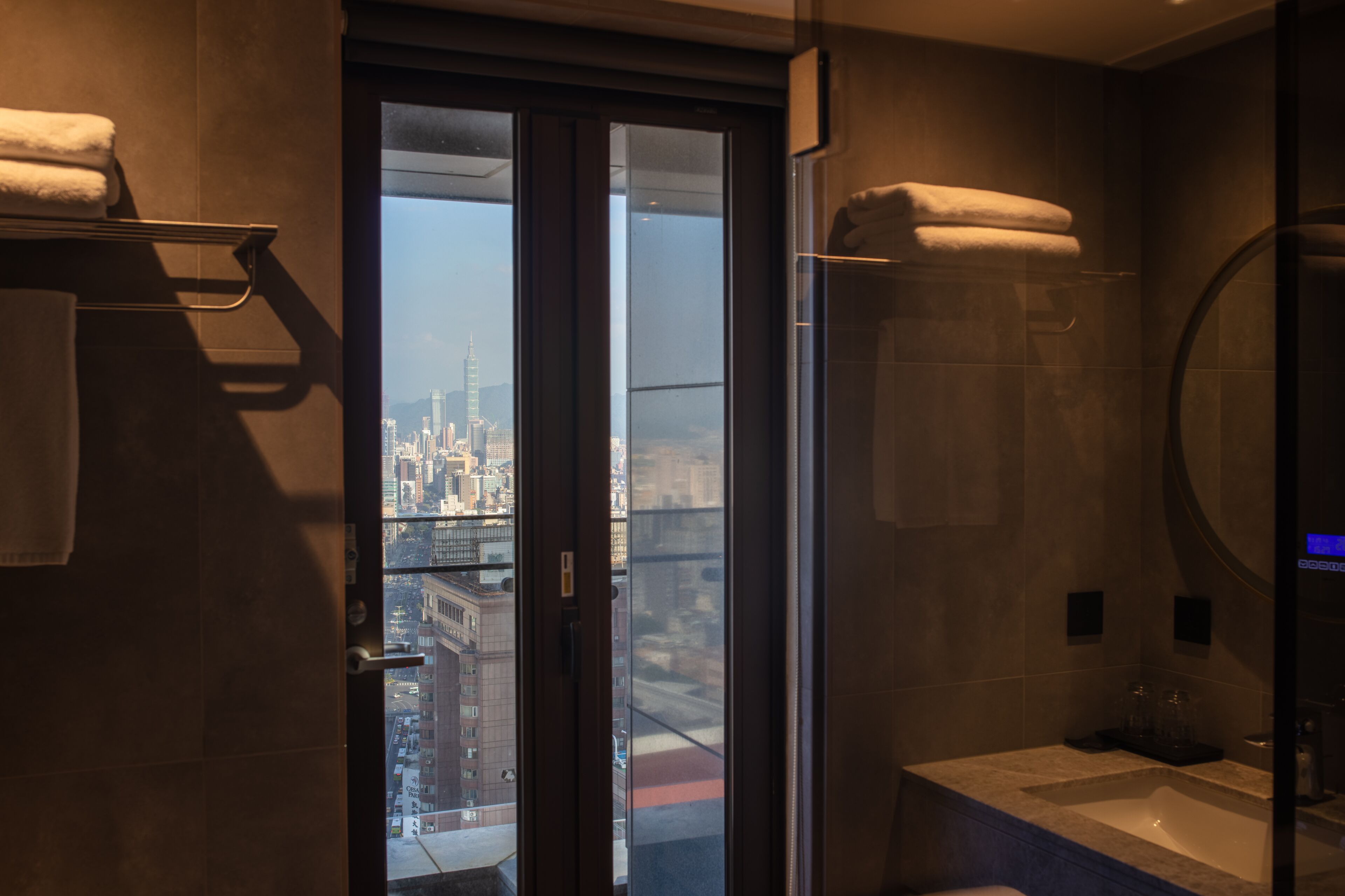 Uma casa de banho moderna com uma banheira grande, uma porta de vidro para uma vista da cidade e toalhas numa prateleira.