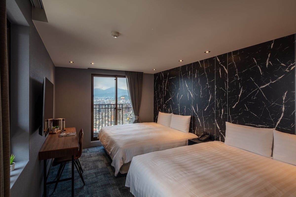 A hotel room with two beds, a desk, a chair, a TV, and a large window with a city view.