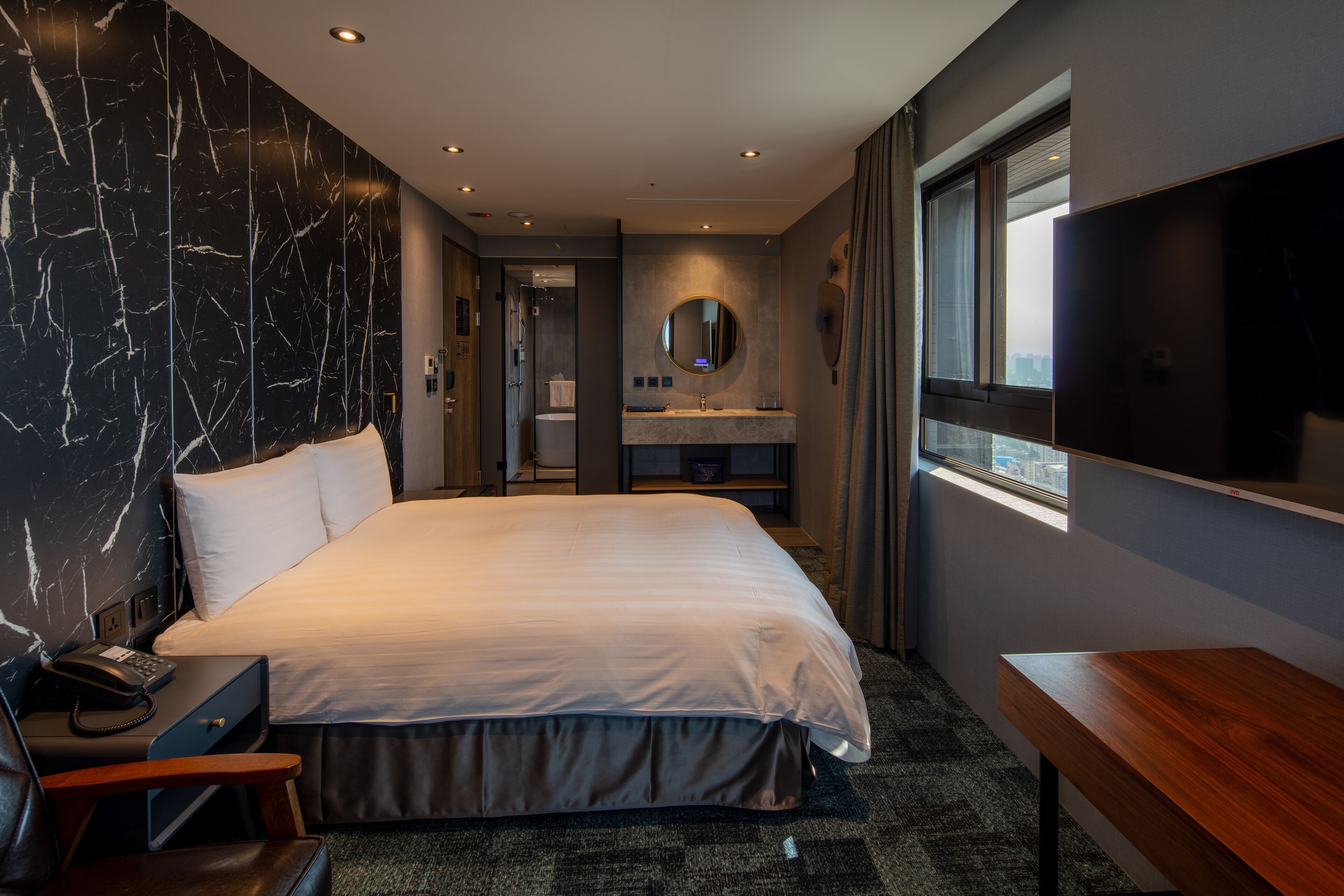 A modern hotel room with a large bed, a bedside table, a desk, a mirror, a window, and a TV.