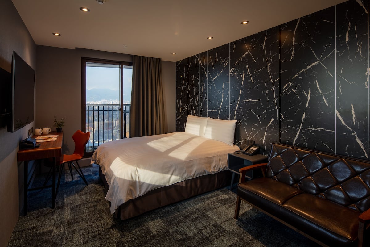 A modern hotel room with a large bed, a leather sofa, a desk with a chair, and a city view through the window.