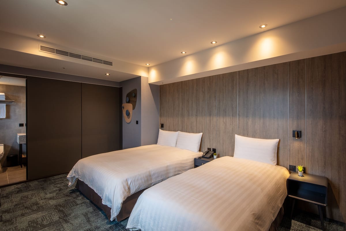 A hotel room with two beds, a wooden accent wall, a small bedside table with a plant, and a modern bathroom with a shower and toilet.
