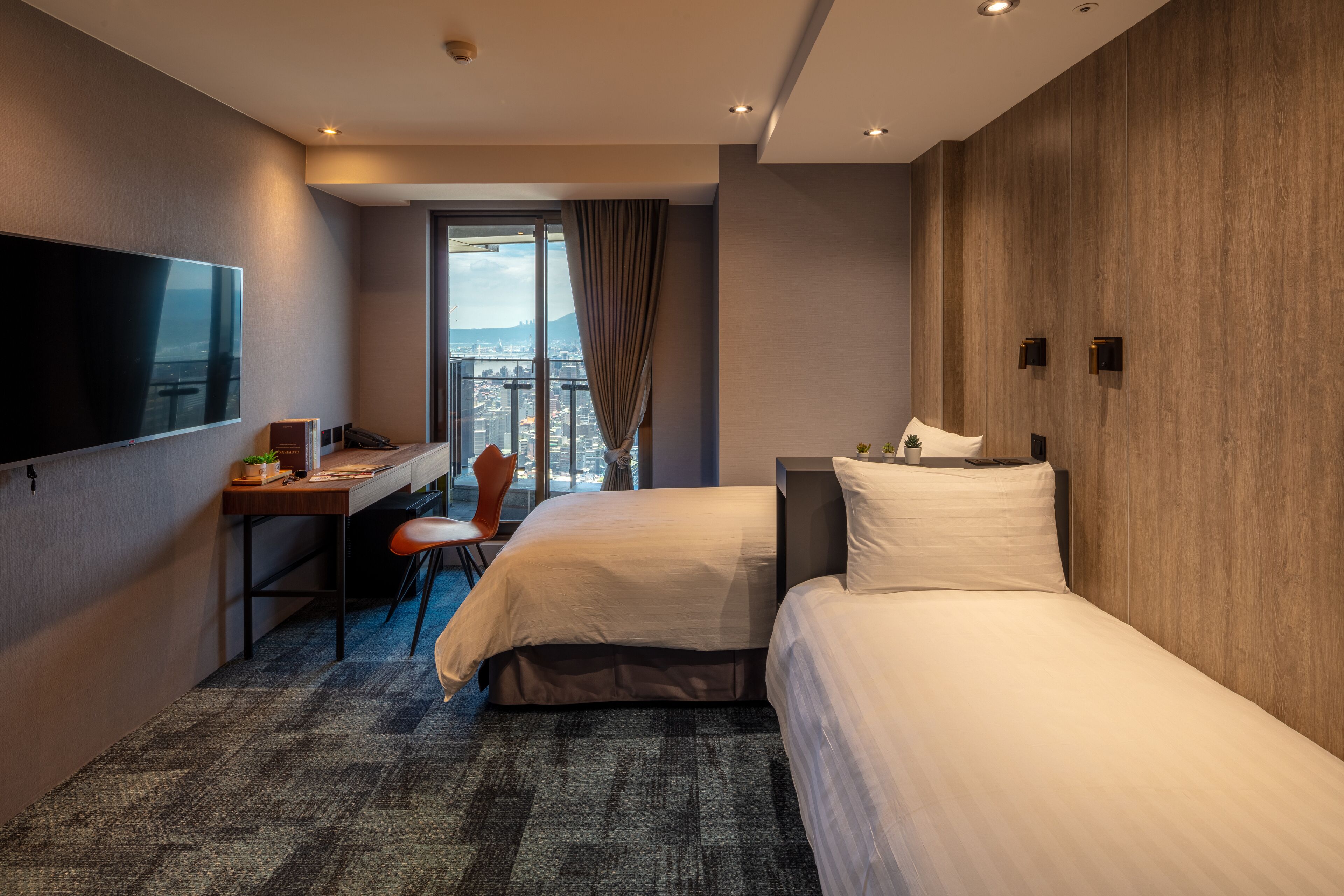 A hotel room with two beds, a desk, a chair, a TV, and a large window with a city view.