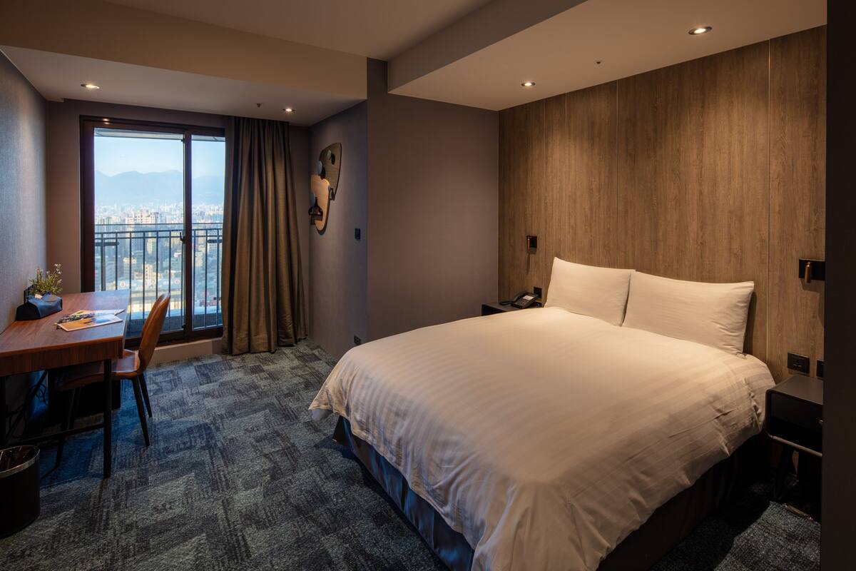 A hotel room with a large bed, a desk with a chair, and a view of a cityscape through a large window.