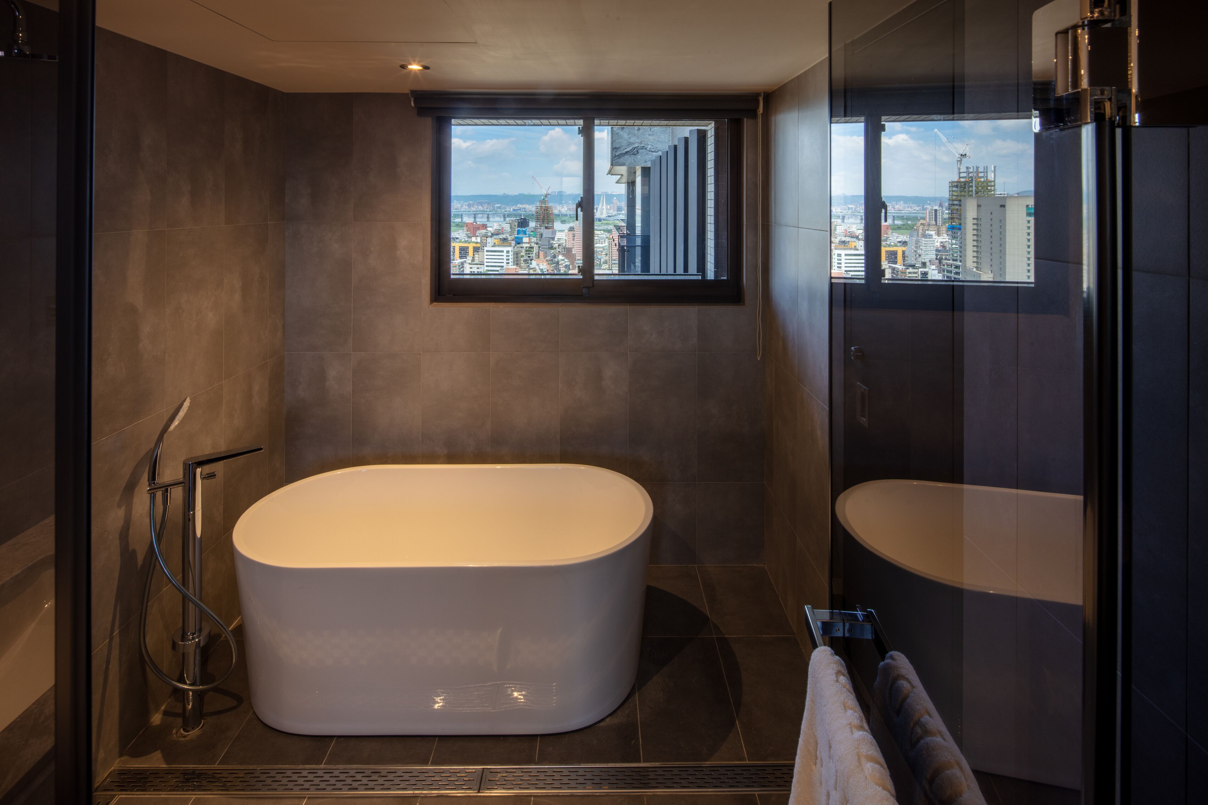 A modern bathroom with a freestanding bathtub, a glass shower enclosure, and a view of the cityscape through a window.