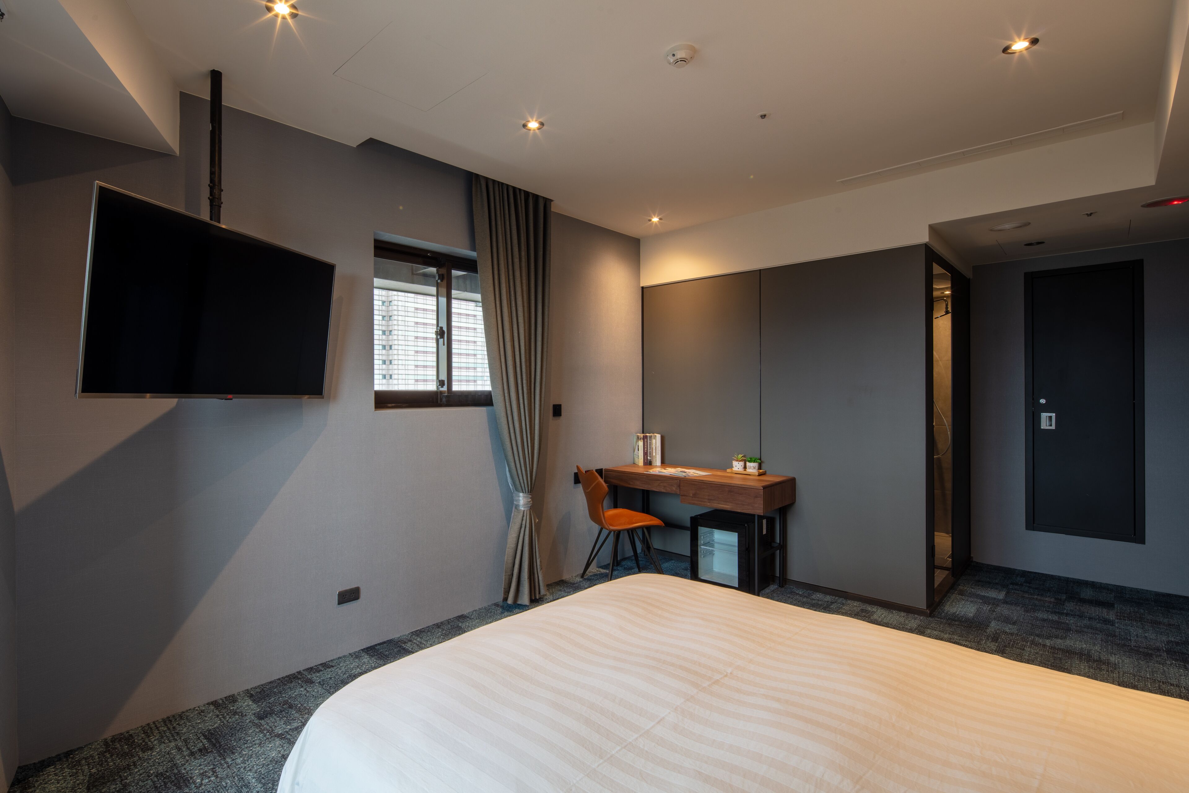 A modern hotel room with a large bed, a desk with a chair, a flat-screen TV mounted on the wall, and a large mirror.