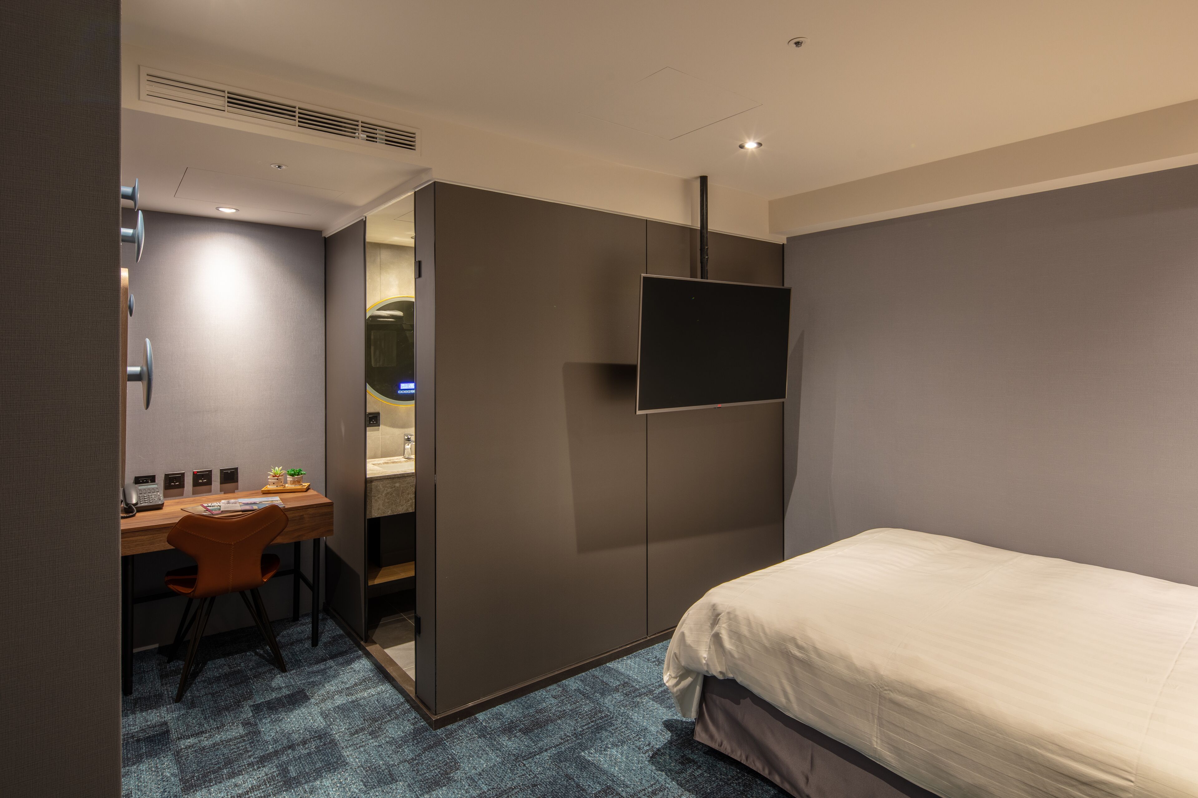 A compact hotel room with a bed, a desk with a chair, a flat-screen TV mounted on the wall, and a small bathroom area visible through a partition.