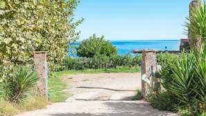 Ein Küstenpfad führt zu einem Steintor mit Blick auf das Meer und einem Gebäude im Hintergrund.