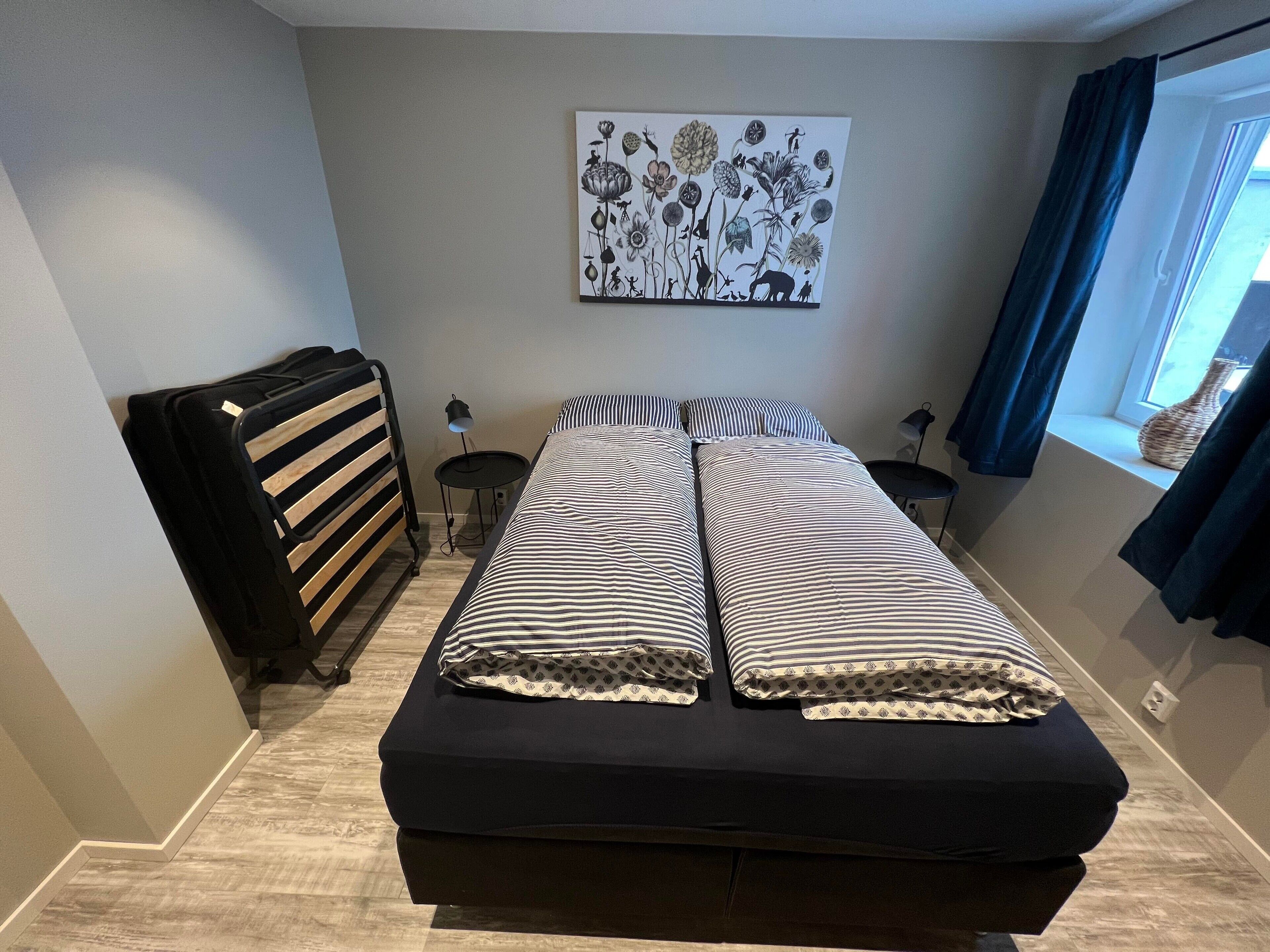 Een slaapkamer met een bed, een radiator, een kleine tafel, een lamp en een raam met gordijnen.