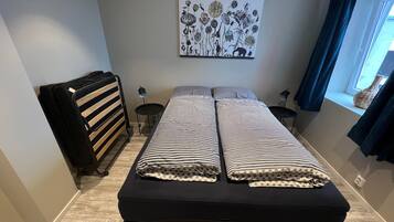 Een slaapkamer met een bed, een radiator, een kleine tafel, een lamp en een raam met gordijnen.