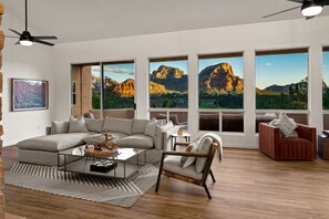 Een moderne woonkamer met een grote modulaire bank, een glazen salontafel en een houten vloer. Er zijn grote ramen met uitzicht op bergen en een plafondventilator.