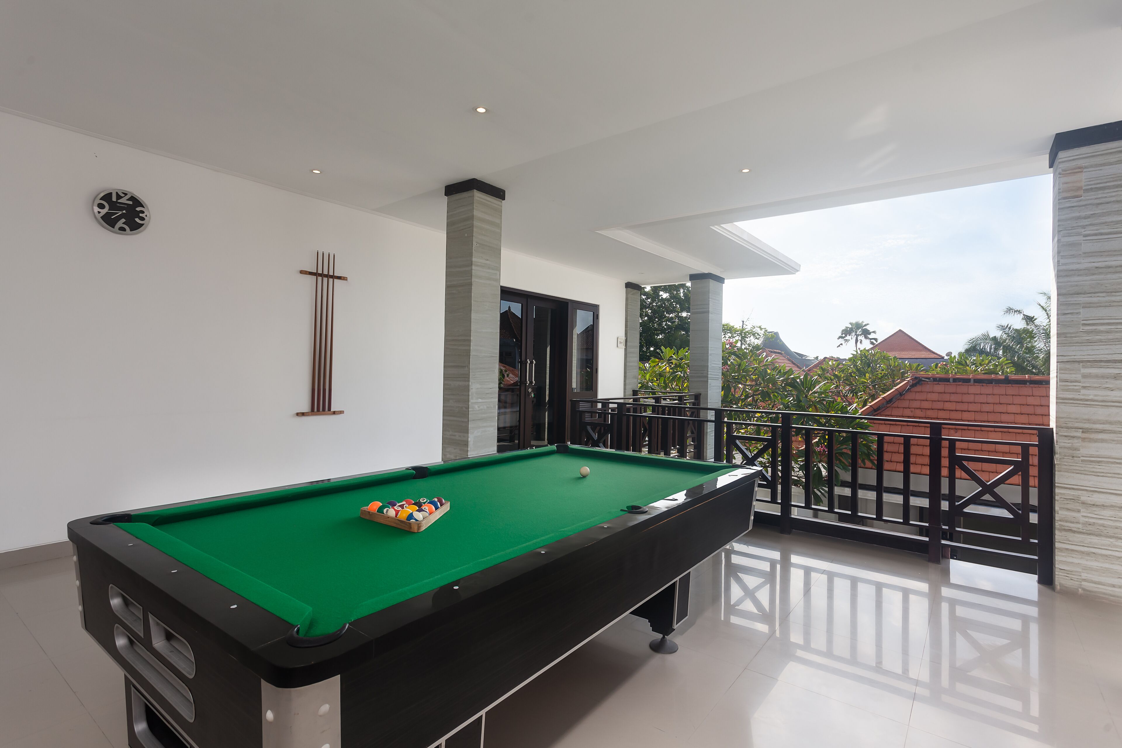 Uma mesa de bilhar em um quarto com varanda, um relógio na parede e vista para edifícios e árvores lá fora.