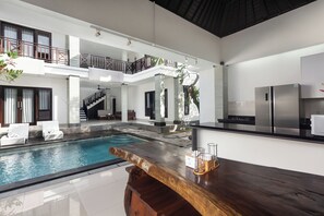 Uma área interna de piscina moderna com um bar de madeira, cadeiras de lounge e uma cozinha com eletrodomésticos de aço inoxidável.