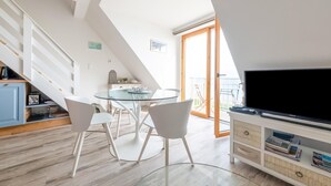 Uma sala de estar moderna com uma mesa de jantar de vidro, cadeiras brancas, uma TV de tela plana e uma escada que leva a uma varanda.