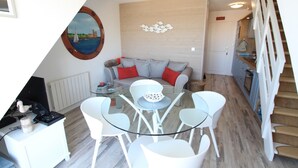 Uma sala de estar moderna com uma mesa de jantar de vidro, cadeiras brancas, um sofá cinza com almofadas vermelhas e um quadro pendurado na parede com a imagem de um veleiro.