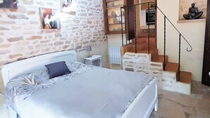 A bedroom with a stone wall, a bed with a white headboard, a nightstand, a wooden staircase, and a shelf with decorative items. - Geräumiges Ferienhaus mit Garten (BREUGNON)