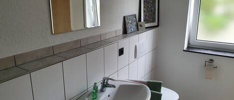 Une salle de bain avec un lavabo blanc, un miroir et des toilettes. Une fenêtre offre une vue sur la verdure à l’extérieur.
