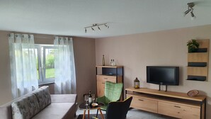 Ett vardagsrum med en soffa, ett matbord med stolar, en TV, ett träskåp och ett fönster med gardiner.